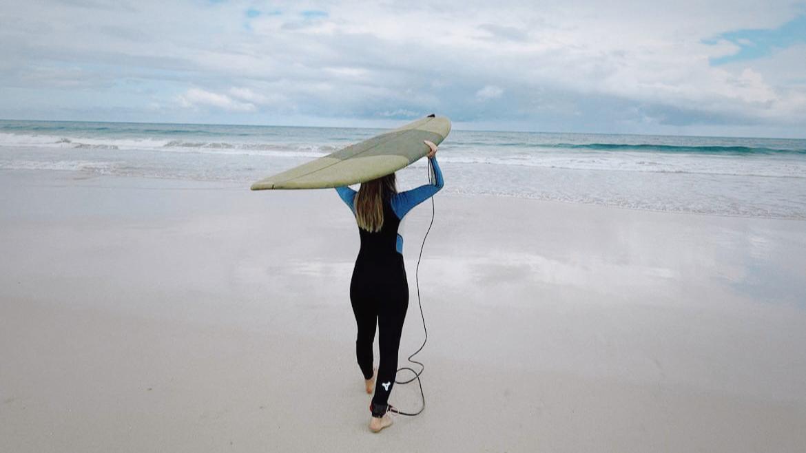 Rebecka bär sin surfbräda på huvudet och går mot stranden, fotografi.